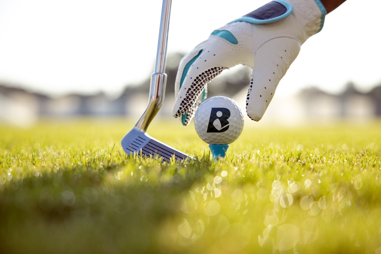 Black People Golf B monogram applied to a golf ball on a tee beside a golf club and gloved golfer’s hand.