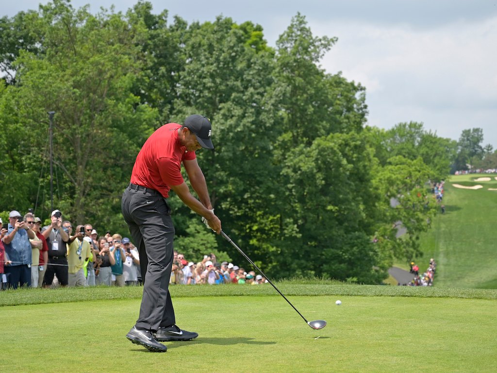 Golfer in a red shirt striking a tee shot on a golf course as spectators watch from behind the tee.