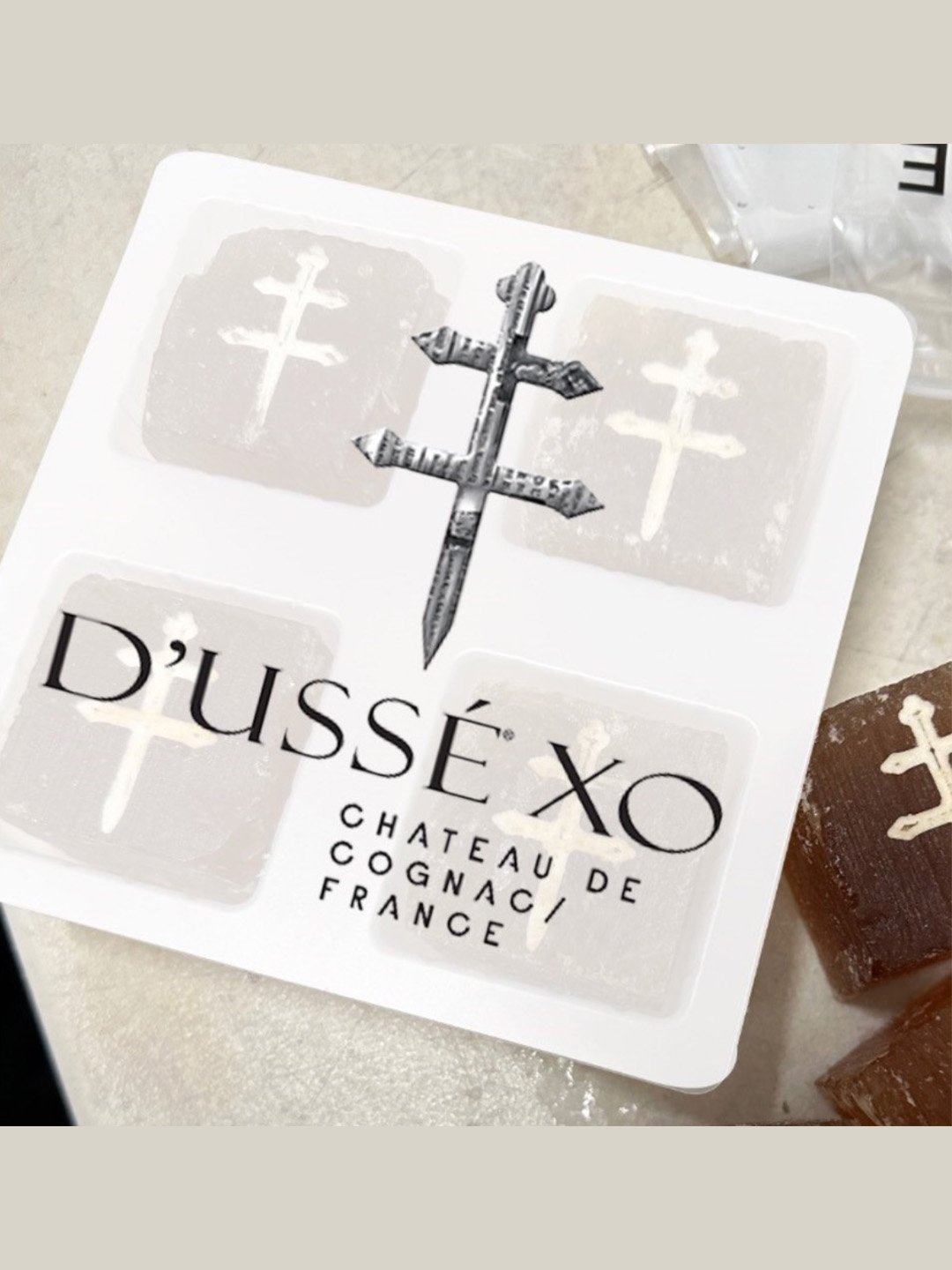 White tray holding four large D’USSÉ XO branded ice cubes with the Cross of Lorraine emblem, beside a brown coffee cube.
