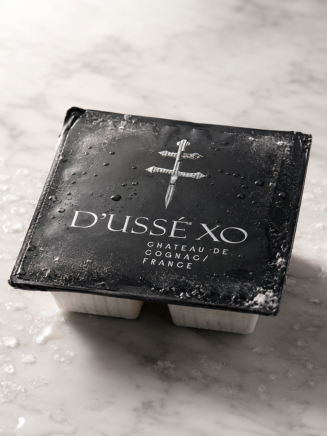 Frozen D’USSÉ XO Coffee Cube package with black branded lid, condensation and frost on a light stone surface.